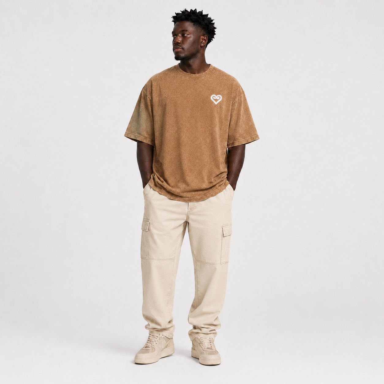 A man modeling a brown acid-wash t-shirt with a small white heart logo on the chest, paired with beige cargo pants and beige sneakers, against a light gray studio background.