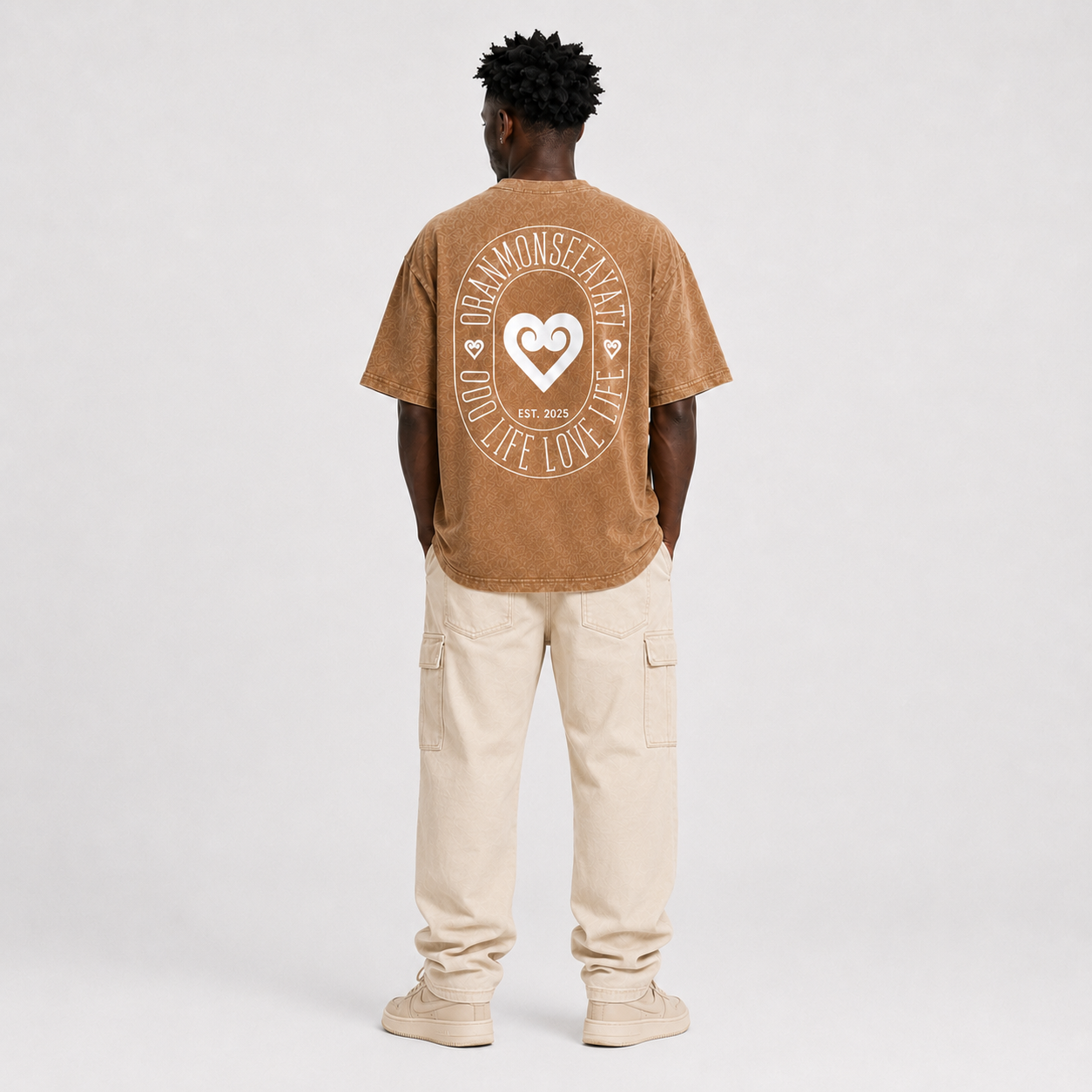 A man viewed from behind wearing a brown t-shirt with a large white circular graphic on the back, paired with beige cargo pants and beige sneakers, against a light gray studio background.