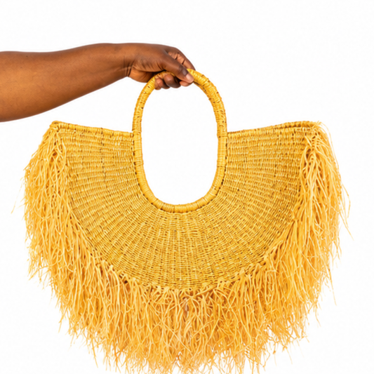 A mustard yellow woven straw tote bag with a single top handle and long fringe, held by a person's hand against a plain white background.