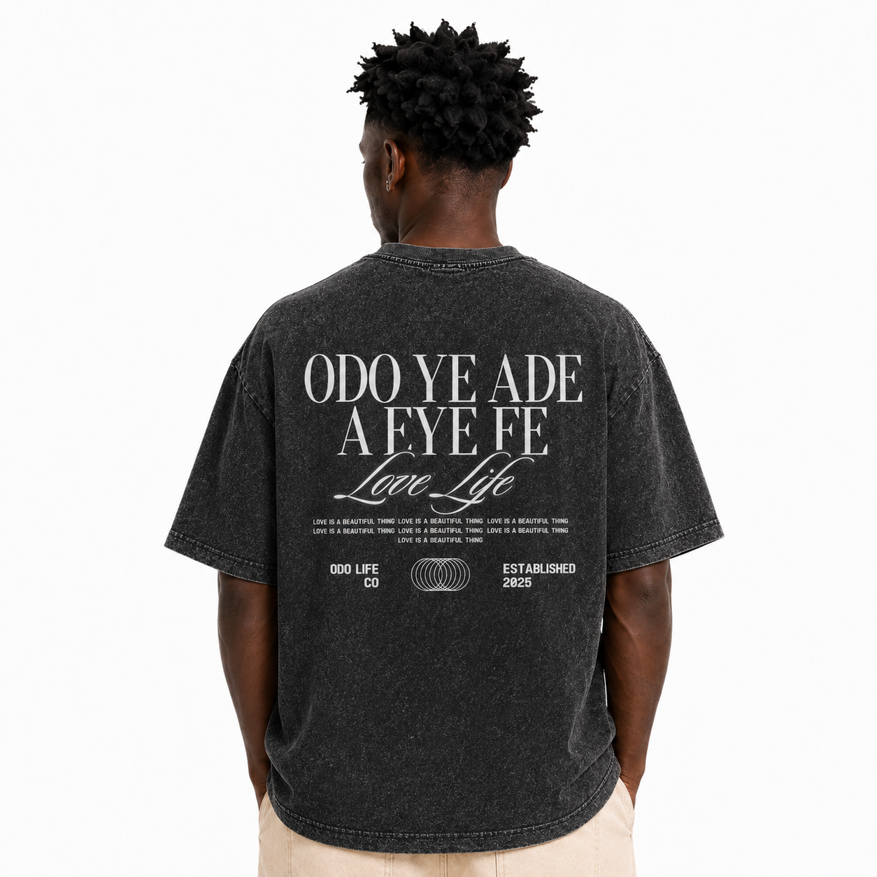 A man viewed from behind wearing a faded black graphic t-shirt with white text on the back reading 'ODO YE ADE A FE FE Love Life' and '000 LIFE CO ESTABLISHED 2025', paired with light beige pants, against a plain white background.
