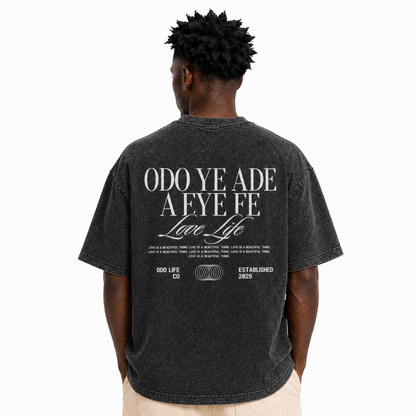 A man viewed from behind wearing a faded black graphic t-shirt with white text on the back reading 'ODO YE ADE A FE FE Love Life' and '000 LIFE CO ESTABLISHED 2025', paired with light beige pants, against a plain white background.