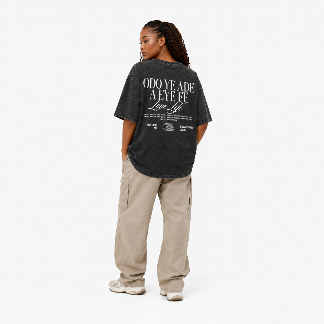 A model shown from the back wearing a dark charcoal gray graphic t-shirt with white text, paired with beige cargo pants and white sneakers, against a plain white studio background.