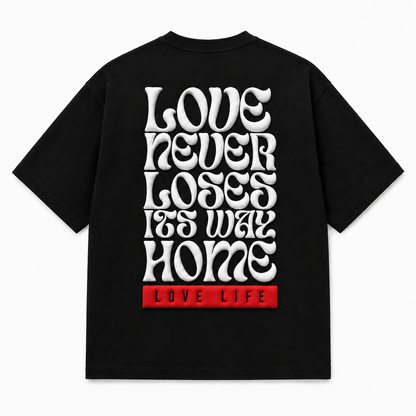 A black short-sleeve t-shirt shown from the back, featuring a large white retro-style text graphic that reads 'LOVE NEVER LOSES ITS WAY HOME' with a red block at the bottom containing the words 'LOVE LIFE'.