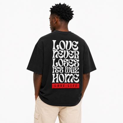 A man viewed from behind wearing a black t-shirt with a large white typographic graphic on the back reading 'LOVE NEVER LOSES ITS WAY HOME' and a red block at the bottom reading 'LOVE LIFE'.