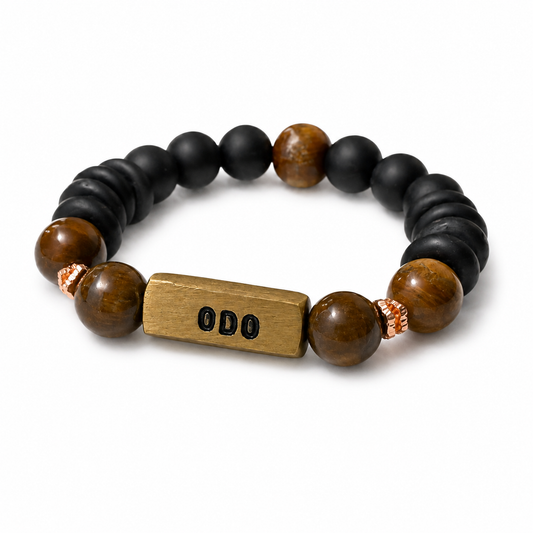 A beaded stretch bracelet featuring matte black round beads, polished brown tiger's eye beads, a rectangular gold-toned metal charm engraved with 'ODO', and small copper spacer beads, shown on a white background.