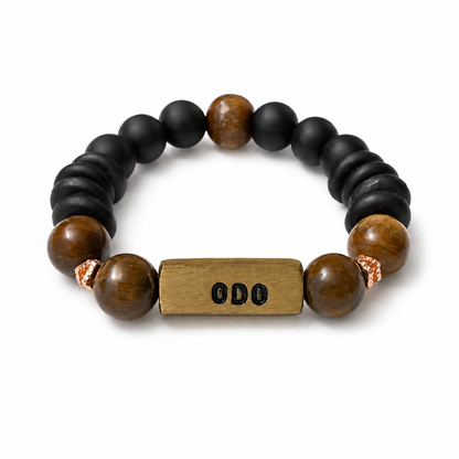 A beaded stretch bracelet featuring matte black round beads, polished brown tiger's eye beads, and a central rectangular brass spacer engraved with 'ODO'.