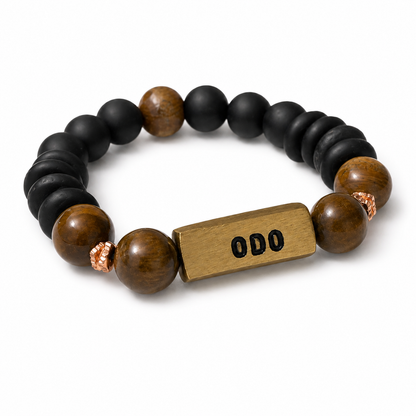 A beaded stretch bracelet featuring matte black round beads, polished brown tiger's eye beads, a rectangular gold-toned metal ID plate engraved with 'ODO', and two small copper-colored spacer beads, shown on a clean white background.
