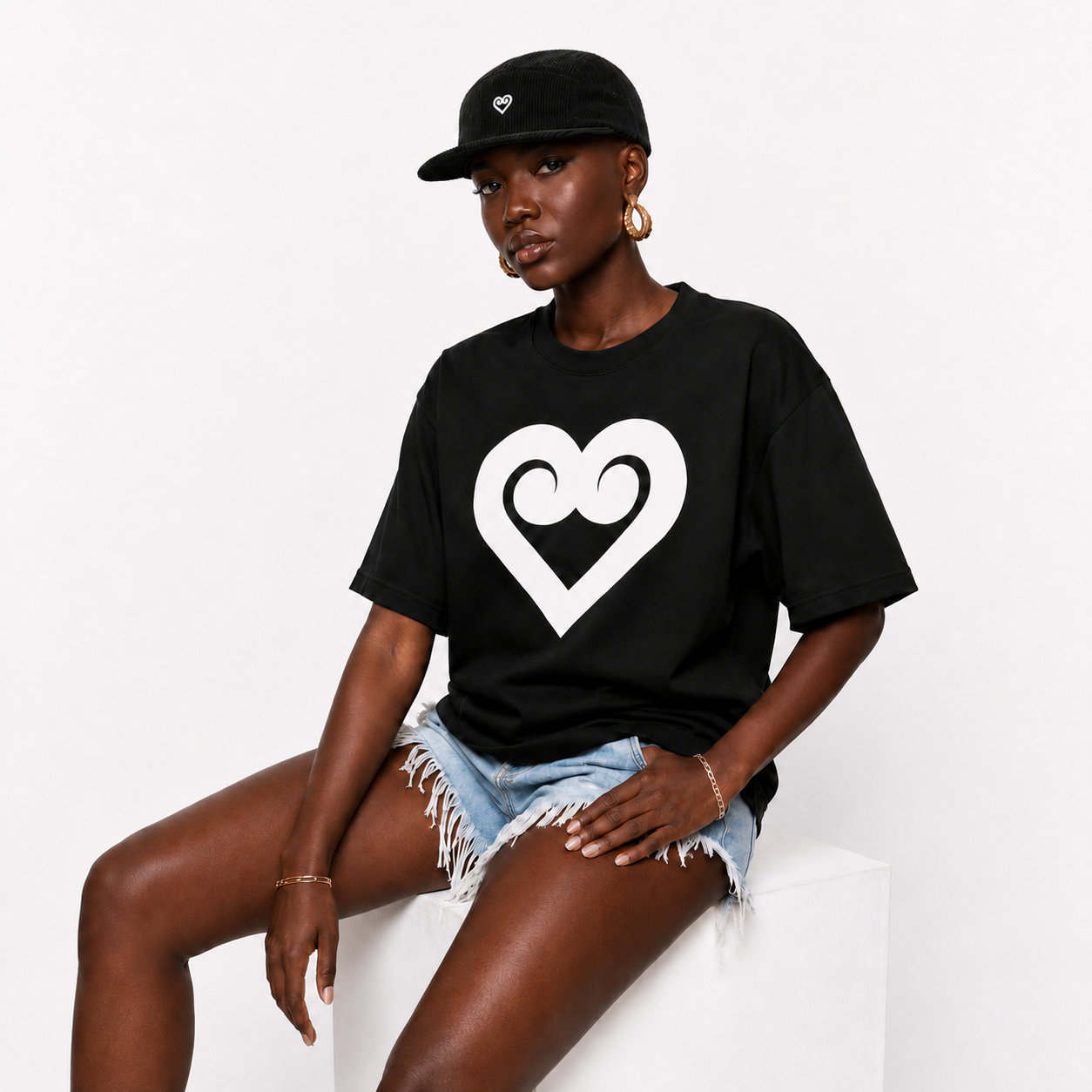 A model wearing a black oversized t-shirt with a large white heart graphic on the front, paired with denim shorts and a black baseball cap, sitting on a white block against a white background.