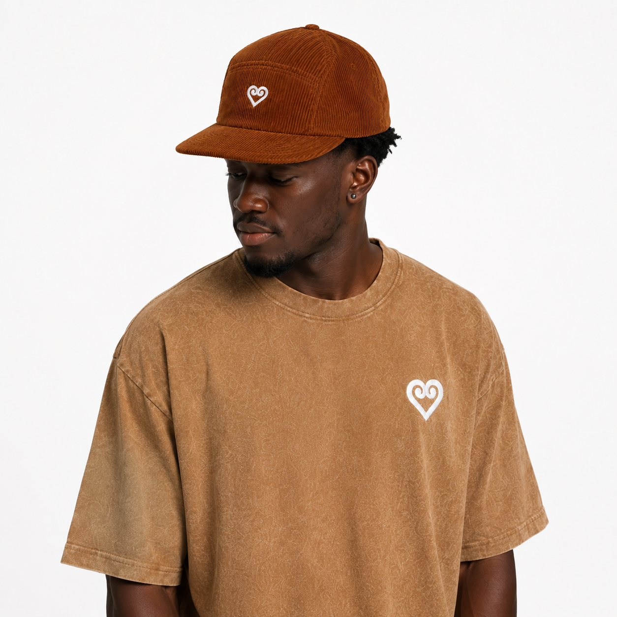 A man wearing a brown corduroy baseball cap and a brown t-shirt, both featuring a small white heart logo on the front, against a plain white background.