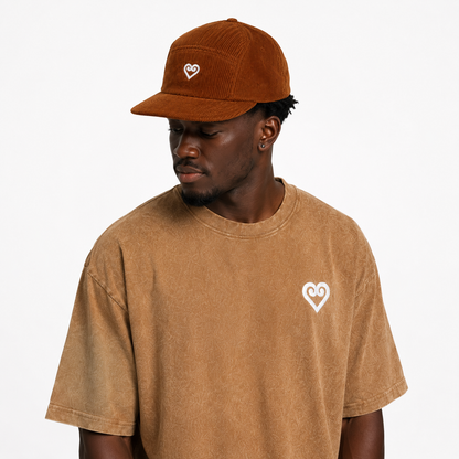 A man wearing a brown corduroy baseball cap and a brown t-shirt, both featuring a small white heart logo on the front, against a plain white background.