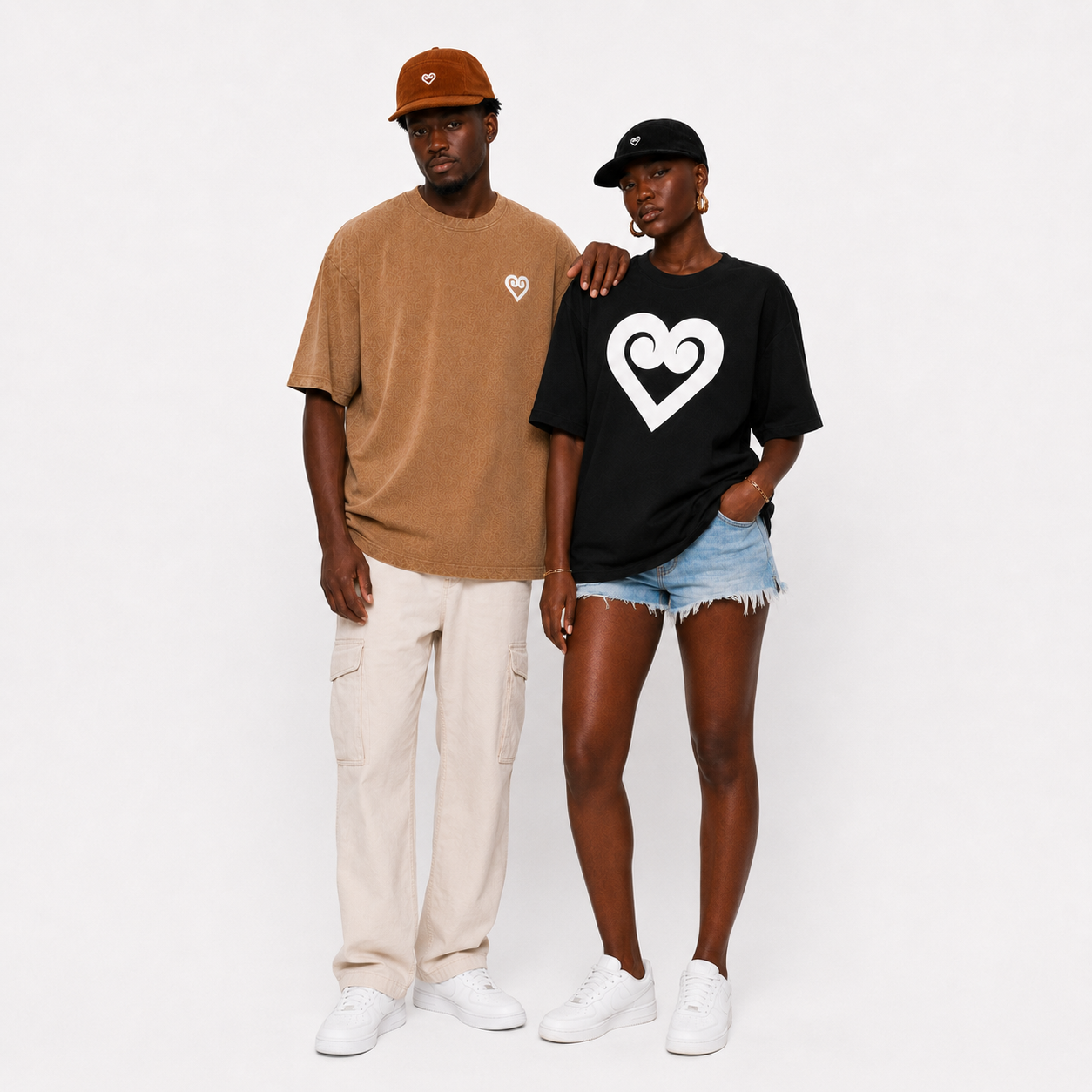A man and a woman modeling oversized graphic t-shirts. The man wears a tan oversized t-shirt with a small white heart logo on the left chest, paired with beige cargo pants, white sneakers, and a brown cap.