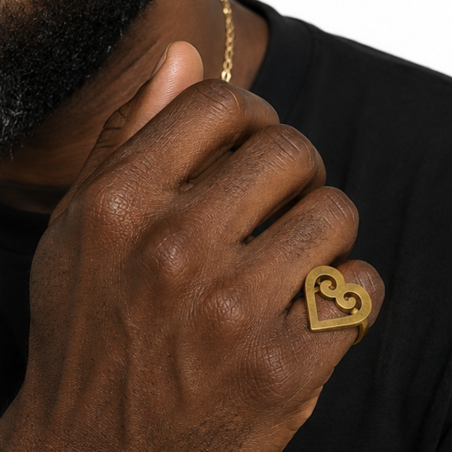 A close-up of a person's hand wearing a gold-colored brass ring with an open heart design featuring a swirl motif inside. The person is wearing a black shirt and a gold chain necklace is partially visible.