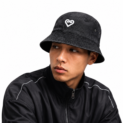 A man wearing a black washed bucket hat with a white heart logo on the front, paired with a black zip-up jacket, photographed against a solid white background.