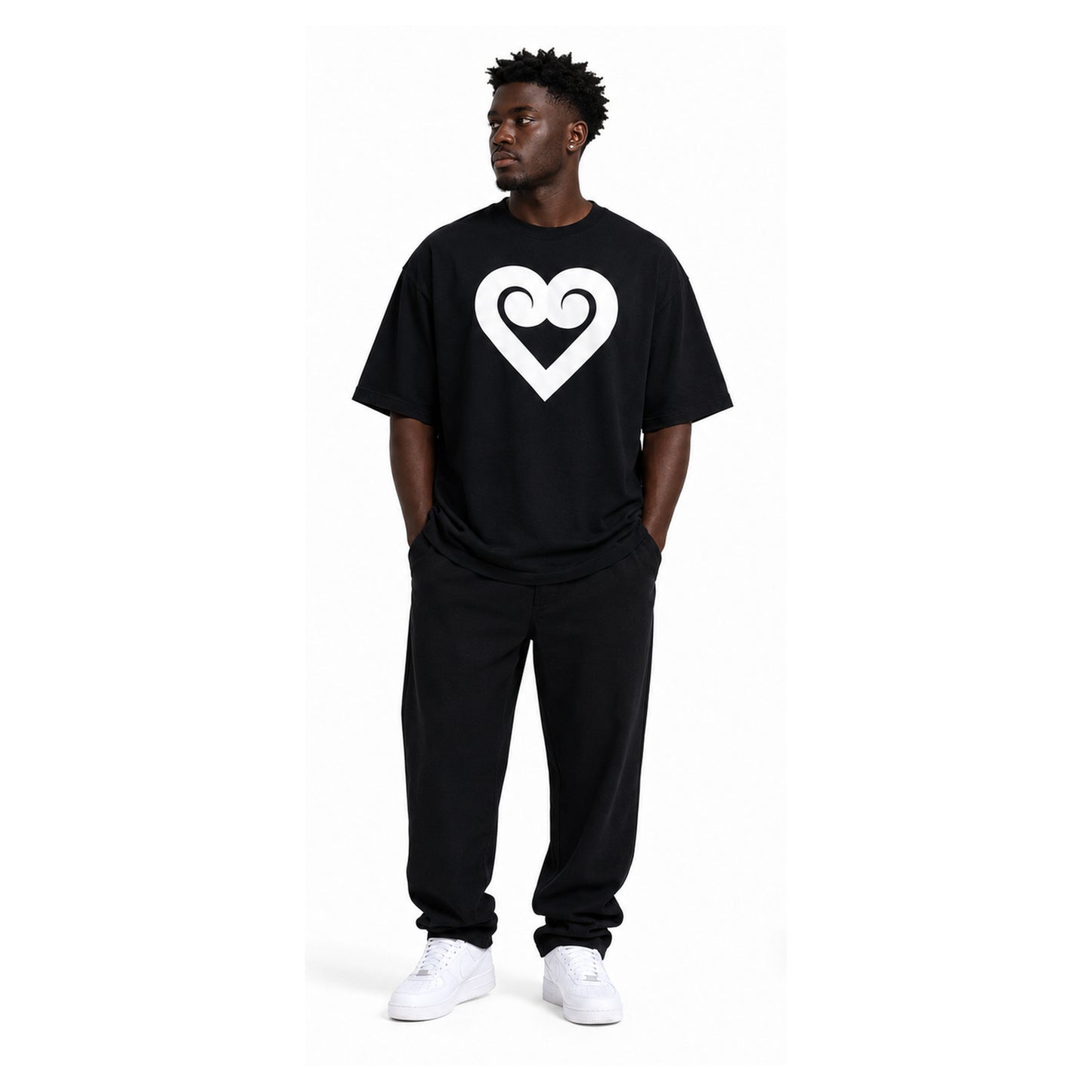 A man wearing a black oversized t-shirt with a white heart logo on the chest, paired with black sweatpants and white sneakers, standing against a plain white background.