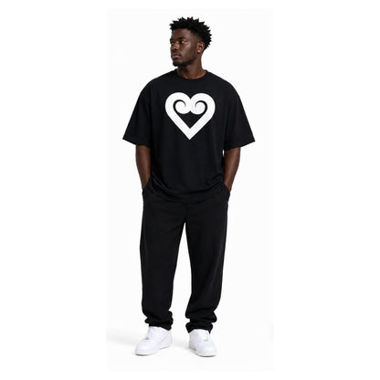 A man wearing a black oversized t-shirt with a white heart logo on the chest, paired with black sweatpants and white sneakers, standing against a plain white background.