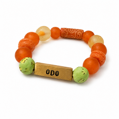 A beaded stretch bracelet featuring orange and light green beads, a carved orange cylindrical bead, and a rectangular gold-toned metal charm stamped with 'ODO'.