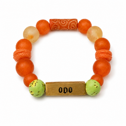 A beaded stretch bracelet featuring round orange and peach beads, a carved orange cylindrical bead, two light beige stone beads, two green marbled beads, and a rectangular gold-toned metal charm engraved with 'ODO'.