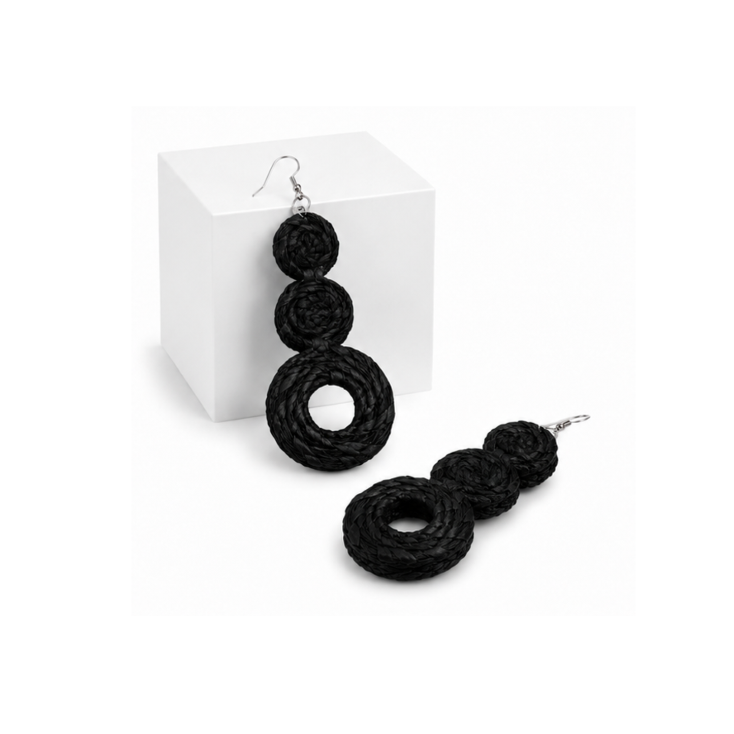 Raffia black braided drop earrings with three stacked spiral rings and silver fishhook wires, displayed on a white cube and a white surface.