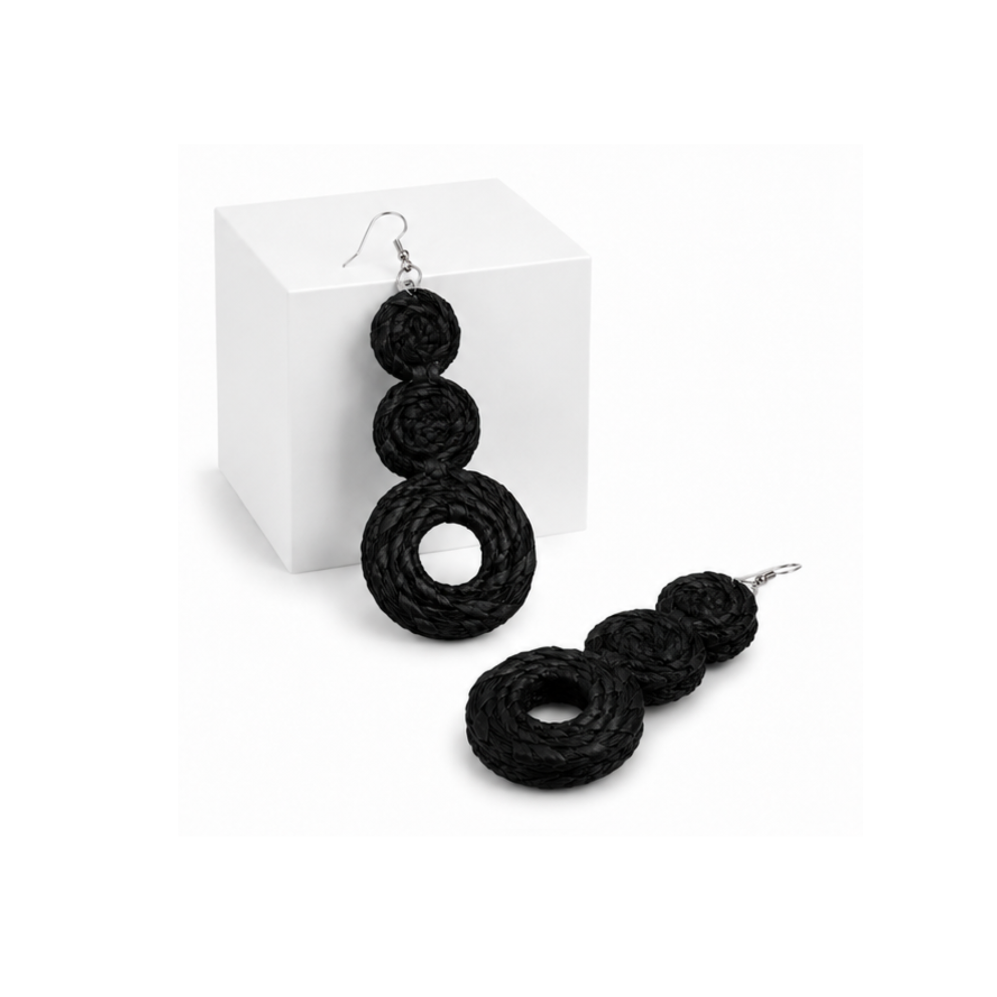 Raffia black braided drop earrings with three stacked spiral rings and silver fishhook wires, displayed on a white cube and a white surface.