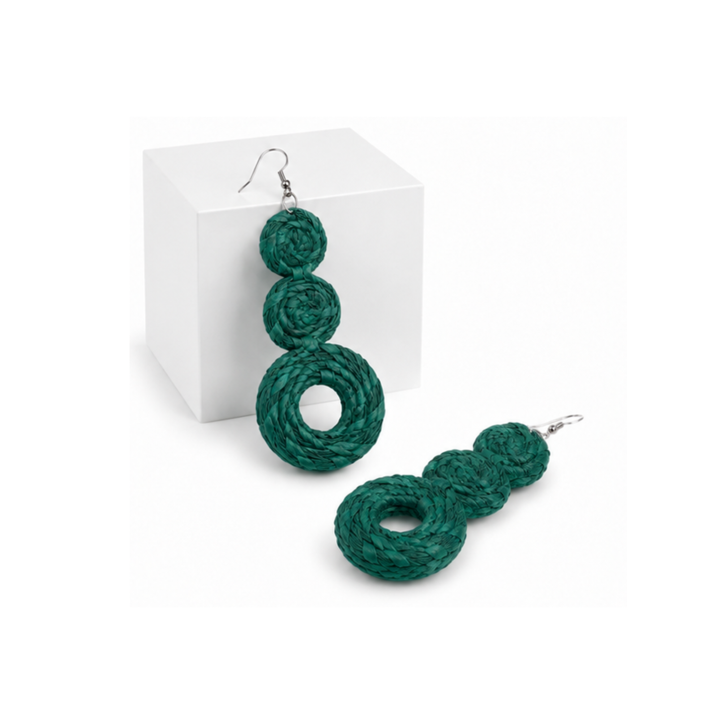 A pair of teal raffia braided rope dangle earrings with silver fishhook wires, displayed on a white cube and a white surface.