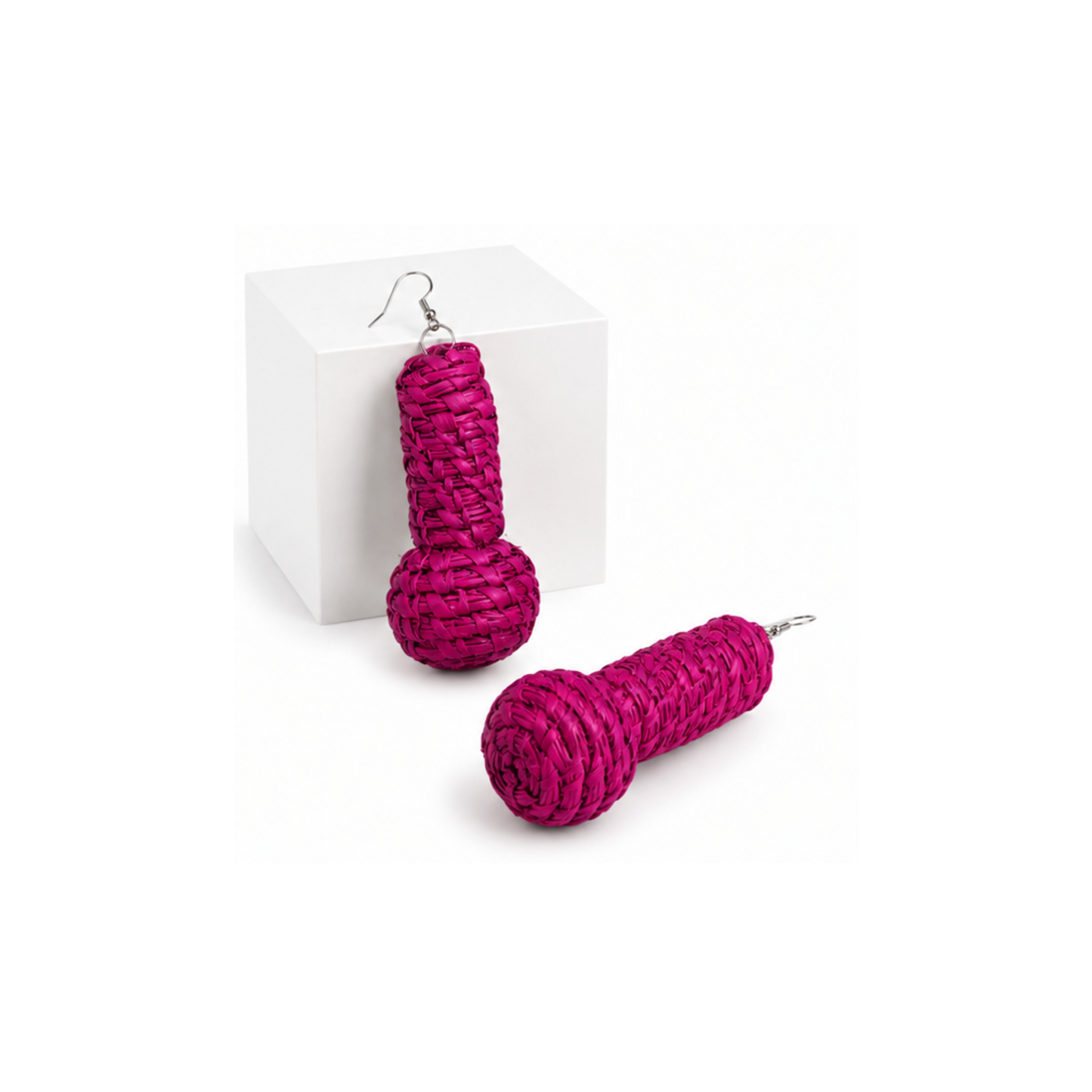 A pair of bright magenta raffia earrings, one resting on a white display block and one on a white surface, both with silver-tone hooks.